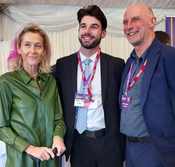 Zac Shaw CACI Digital Experience Accessibility Lead stands smiling wearing a smart suit between a pink-haired Baroness Martha Lane Fox of Soho who is holding her cane and Mark Walker of AbilityNet