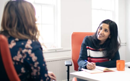 Photo of Meera interviewing a client at a desk. She is writing notes in her notepad and smiling as she listens to the client.